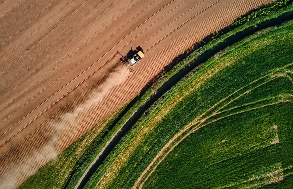 Birds eye view of a field, split between dirt and grass for farming, with a tractor moving diagonally mid-picture
